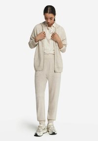 Beige hoodie med frontdragkedja, ribbade ärmslut och två fickor över en randig skjorta och matchande beige byxor. Silverfärgade sneakers fulländar looken.