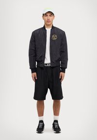 Svart bomberjacka med en logotyp på bröstet, vit t-shirt med rund hals, svarta shorts och svarta sneakers med vita detaljer; ljus keps.