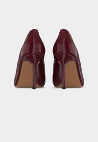 Ζευγάρι γυαλιστερών ψηλοτάκουνων παπουτσιών σκούρου κόκκινου χρώματος, εμφανιζόμενα από πίσω, με λεπτά τακούνια και διακοσμητικό με χρυσό λογότυπο κοντά στο πάνω μέρος.