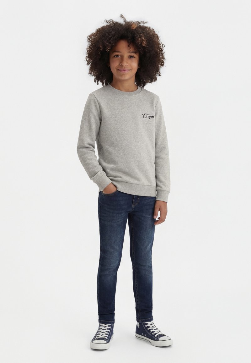 Kind mit lockigem Haar, das einen grauen Sweatshirt, dunkelblaue Jeans und marineblaue High-Top-Sneaker trägt und mit einer Hand in der Tasche steht.