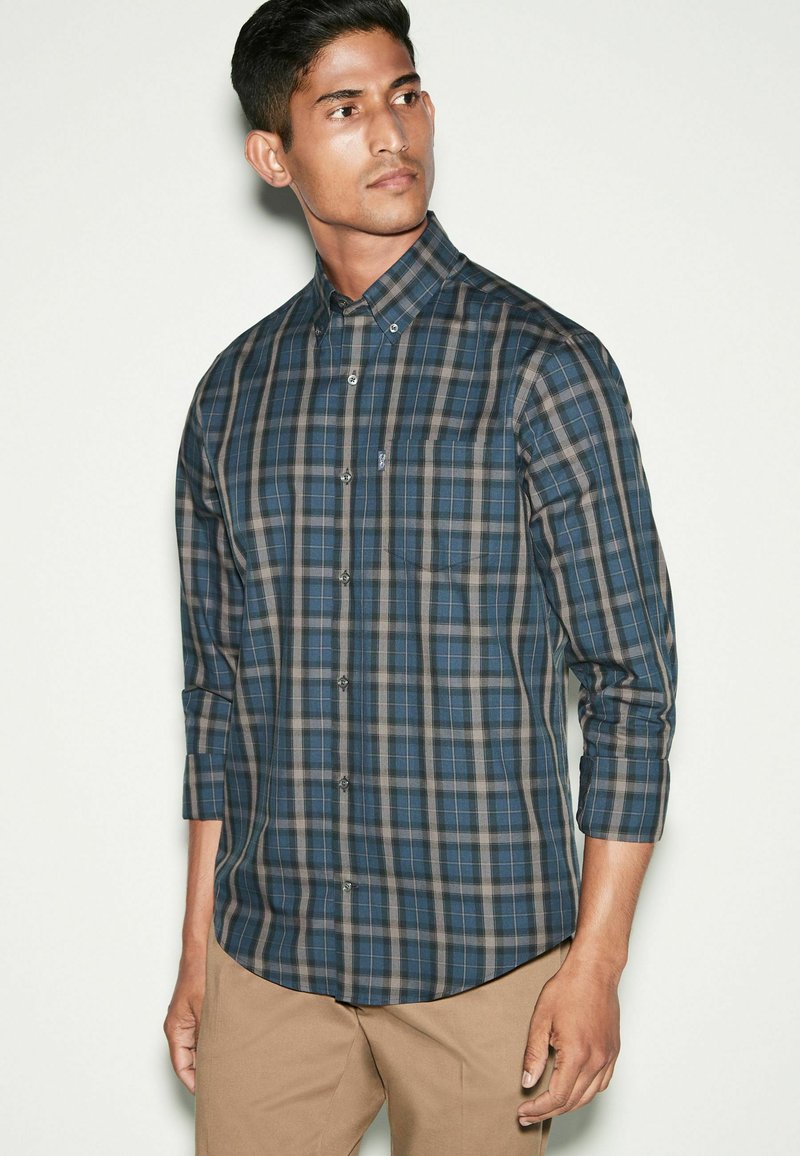 Camisa de cuadros de botones en azul oscuro, gris y beige. Presenta mangas largas, un cuello y un bolsillo en el lado izquierdo del pecho. Textura de tela suave.