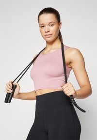 Svijetloružičasti rebrasti crop top s okruglim izrezom uparen s crnim tajicama visokog struka; drži crnu skakaču sa teksturiranim ručkama.