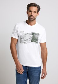 Man med kort brunt hår som bär en vit grafisk T-shirt med berglinjer och text, tillsammans med blå jeans, står mot en ljusgrå bakgrund.