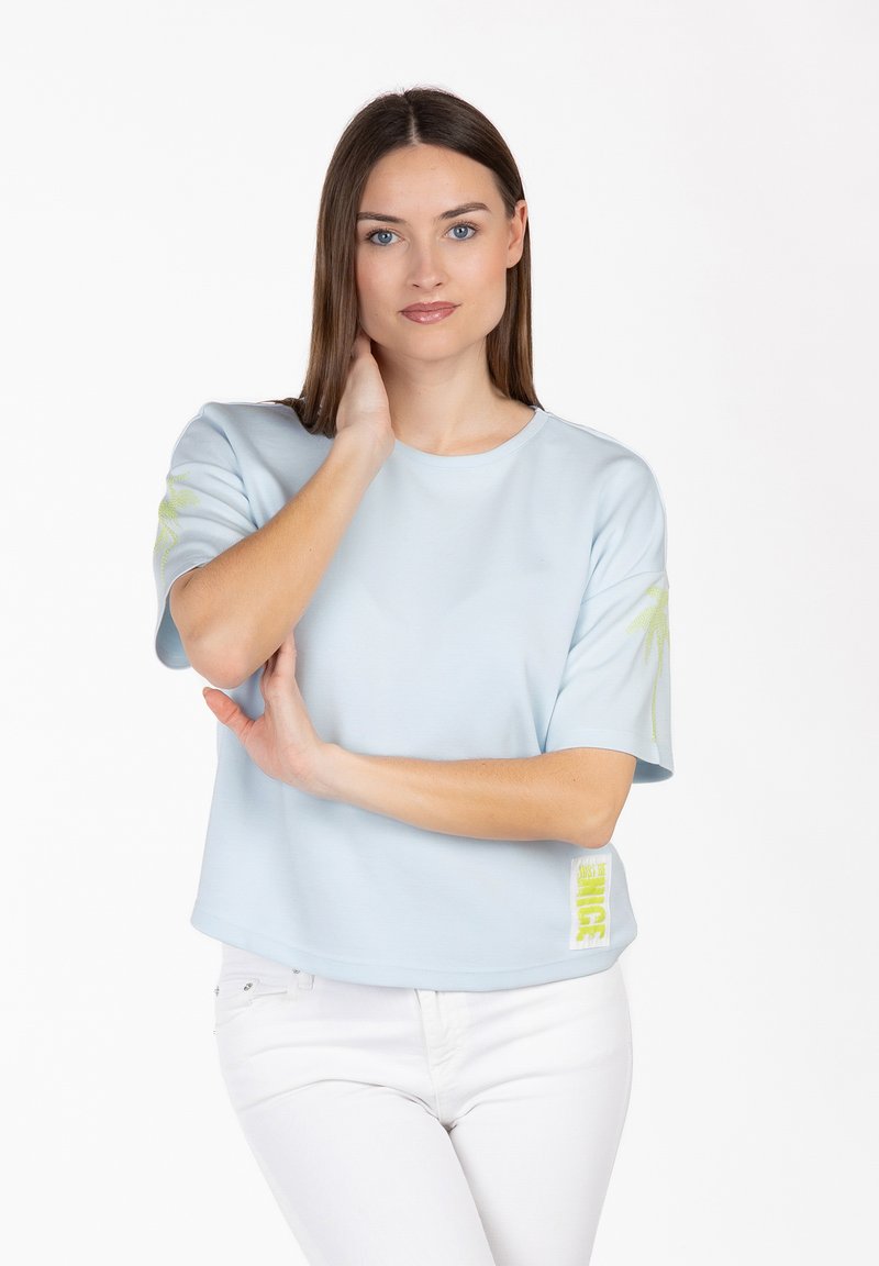 Mujer con cabello castaño liso, vistiendo una camiseta azul claro de manga corta y pantalones blancos, de pie con una mano en el cuello.