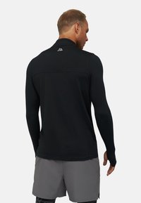 Schwarzes, langärmliges Sportoberteil mit einem Viertel-Reißverschluss, glatter Textur und reflektierendem Akzent auf dem Rücken, kombiniert mit grauen Shorts.
