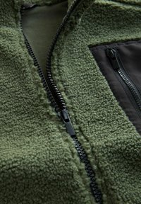 Green fleece jacket featuring a textured surface, black zipper, and a solid black panel. Designed with a full-length zipper and soft lining.