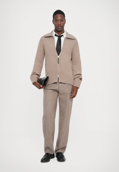 Cardigan marron à côtes avec fermeture éclair à l'avant, col léger, porté sur une chemise blanche et une cravate noire, associé à un pantalon texturé gris et des chaussures noires.