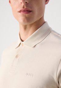 Bežové polo tričko s límečkem, má dvě knoflíky a vyražené logo na levé hrudi. Texturovaná látka a minimální detaily.
