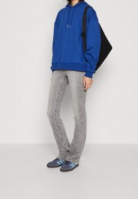 Felpa blu con cappuccio e tasca frontale, jeans slim-fit grigi e sneakers blu con strisce nere e blu, abbinate a una borsa a tracolla nera.