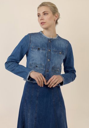 Frau mit blondem Haar trägt eine geknöpfte blaue Jeansjacke mit ausgefransten Kanten und einen passenden Jeansrock und posiert vor hellem Hintergrund.