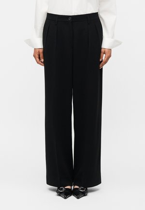 Femeie îmbrăcată cu pantaloni negri cu talie largă și pliuri, nasture în față, buzunare laterale, cămașă albă cu mâneci lungi și pantofi negri cu vârf ascuțit și catarame.