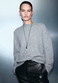 Suéter gris de punto grueso con un patrón texturizado, combinado con pantalones de cuero negros. Presenta un collar largo negro con un colgante.