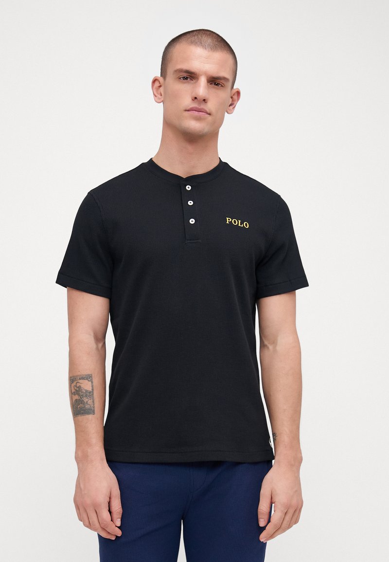 Pólo de algodão preto com mangas curtas, tecido texturizado, pala com três botões e um logótipo amarelo "POLO" no peito.