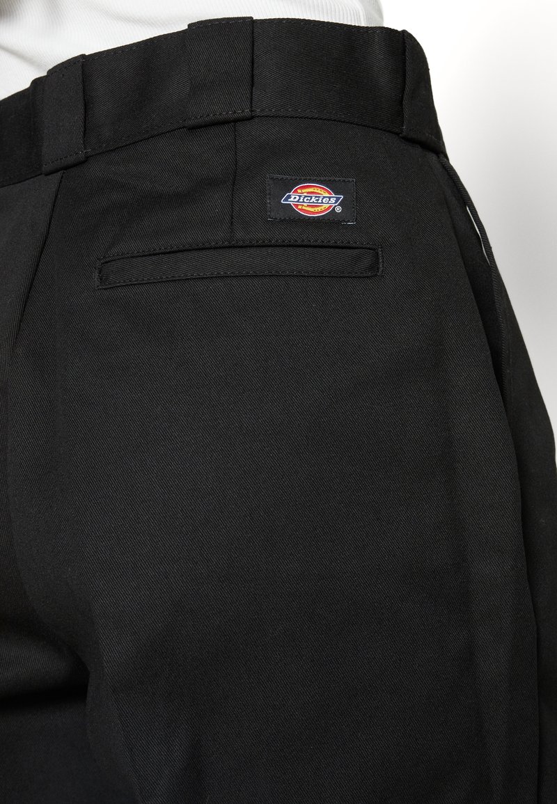 Pantalons de travail noirs en tissu durable, avec une poche arrière ornée d'une étiquette rectangulaire Dickies. Lignes épurées et coupe ajustée.