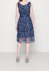 Robe marine avec broderies florales, design sans manches, jupe translucide, taille cintrée, et chaussures à talons hauts bleu clair.