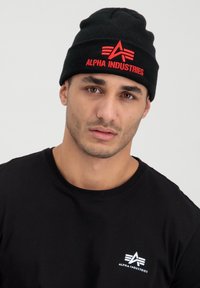 Black knit beanie with prominent red "ALPHA INDUSTRIES" logo and graphic. Worn with a round-neck black t-shirt featuring a white emblem.
