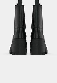 Paire de bottes en cuir noires à talons hauts avec semelles épaisses vues de dos sur un fond clair uni.
