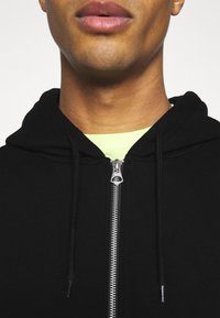Felpa nera con zip e cappuccio regolabile, zip argentata e t-shirt verde lime visibile nel colletto, texture del tessuto liscia.