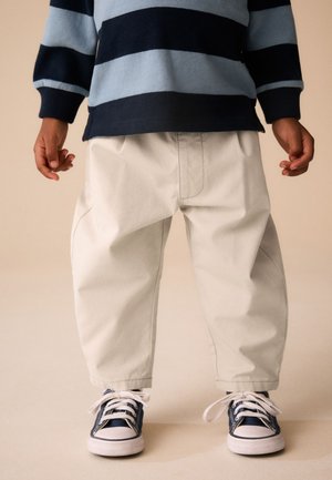 Barn iført løse lyse bukser, marineblå og lyseblå stribet sweater og marineblå sneakers med hvide snørebånd, stående på en beige gulv.