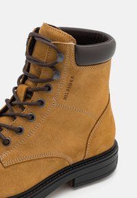 Bottine en suede tan avec des accents en cuir brun, bout rond et lacets marron. Présente des coutures décoratives et une semelle plate noire.