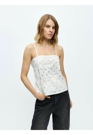 Young woman with shoulder-length blonde hair wearing a white spaghetti strap top with black dots and black pants, posing with one hand in pocket.