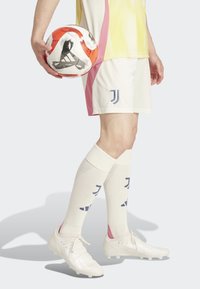 Valkoinen jalkapallopaita, jossa on vaaleanpunaisia ja keltaisia yksityiskohtia, valkoiset shortsit, valkoiset nappulakengät ja polvipituiset sukat, joissa on laivastonsinisiä kuvioita, pitäen punavalkoista jalkapallon.