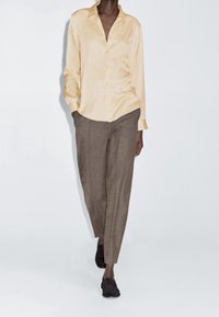 Chemise boutonnée en satin jaune clair associée à un pantalon à carreaux marron et à des chaussures noires texturées, sur un fond blanc uni.