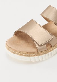 Metallinruskea sandaali, jossa on kaksi leveää remmiä, teksturoitu pinta ja vaaleanvärinen espadrillepohja, jossa on uurrettu reuna.