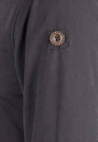 Svart tygjacka med en texturerad lapppatch, brun knapprad och ett präglat logotyp. Tyget verkar hållbart och slätt.