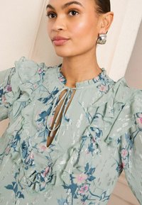 Žalia gėlėta bluzonė su volantais, giliu iškirpimu ir užrišimu. Medžiagoje yra sidabrinių akcentų ir mėlyni gėlių raštai.