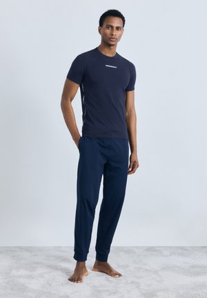 Noor mees seistes paljajalu vaibal, seljas tumesinine Emporio Armani t-särk ja sobivad dressipüksid, käed taskutes, neutraalne taust.