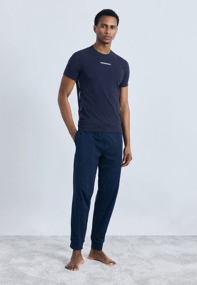 Joven de pie descalzo sobre una alfombra, lleva una camiseta de Emporio Armani azul marino y pantalones de chándal a juego, con las manos en los bolsillos, fondo neutro.