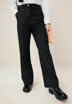 Vrouw draagt zwarte rechte broek met hoge taille, wit overhemd, houdt een beige notitieboek vast en draagt zwarte glanzende veterschoenen.