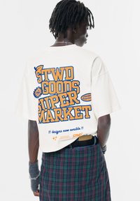 Biała koszulka krótki rękaw z niebieską i pomarańczową typografią na plecach, z napisem "STWD GOODS SUPERMARKET" oraz dodatkowymi graficznymi akcentami.