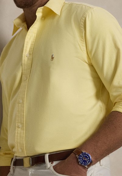 Bărbat purtând o cămașă galbenă cu nasturi, cu un mic logo brodat, pantaloni albi, curea maro și ceas de mână cu cadran albastru și curea maro.