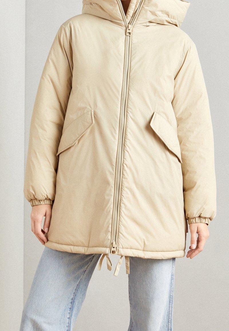 Veste matelassée beige à capuche avec fermeture éclair frontale, deux poches à rabat inclinées, poignets élastiques et ourlet à cordon de serrage, portée sur un jean délavé clair.
