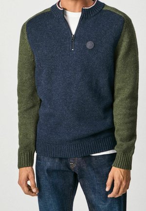 Mann trägt blauen und grünen Strickpullover mit Halb-Reißverschluss-Kragen über weißem Hemd und blauer Jeans, Hände ruhen an den Seiten.