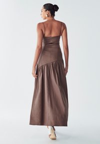 Maxikleid aus braunem Stoff mit einem taillierten Oberteil und einem ausgestellt, rüschengesäumten Rock. Zarte Spaghettiträger und ein dezenter Reißverschluss auf der Rückseite.