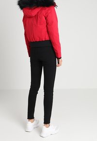 Červená zateplená bunda s černým kožíškem na kapuci, zkrácený střih, nošena s černými úzkými džíny a bílými teniskami. Hladká textura.