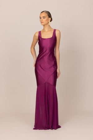 Femme debout portant une robe en satin violette sans manches et ajustée, avec une jupe inférieure transparente, et des boucles d'oreilles pendantes dorées à motif floral.