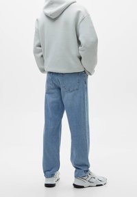 Světle šedá mikina s kapucí, volné modré džíny a bílé tenisky. Džíny mají klasický pětikaprový design a vybledlou texturu.