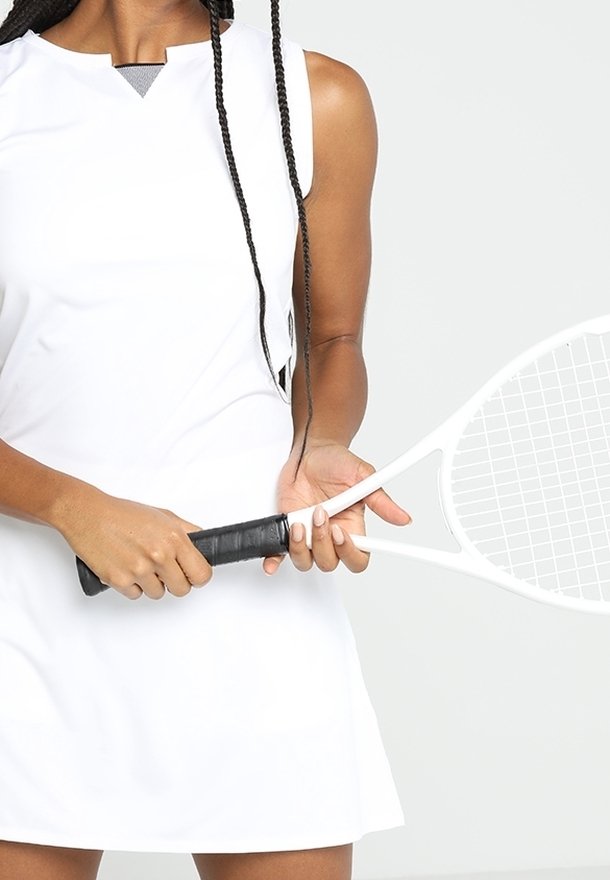 Femme en robe de tennis blanche tenant une raquette de tennis blanche à deux mains sur un fond clair uni.