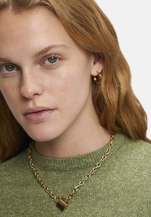 Collier en or avec un pendentif en forme de cadenas, porté sur un pull en maille verte ; le modèle a la peau claire et de longs cheveux ondulés.