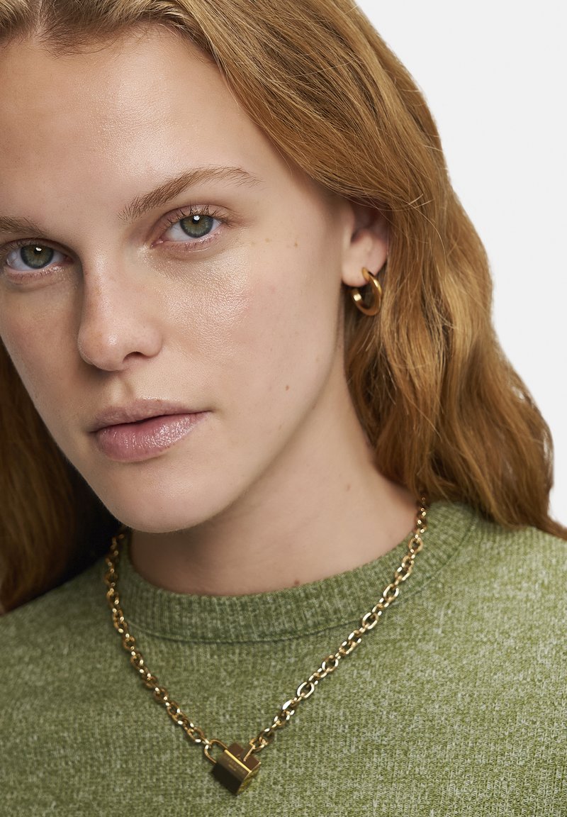 Gouden ketting met een hangslot hanger, gedragen over een groene textuurtrui; model heeft een lichte huid en lang, golvend haar.