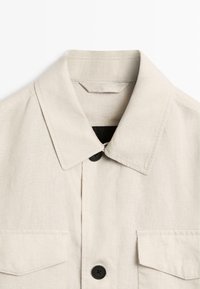 Beige Hemd mit Kragen, zwei aufgesetzten Taschen vorne und sichtbaren schwarzen Knöpfen auf weißem einfarbigem Hintergrund.