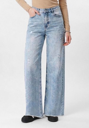 Osoba nosi svetlomodré roztrhané rifle s širokými nohavicami, béžový dlhý rukáv, čierne topánky a strieborné náramky na zápästí.