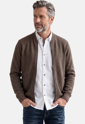 Mann mit grauen Haaren und Bart, der einen braunen Pullover mit Reißverschluss über einem weißen Hemd und blauen Jeans trägt, Hände in den Taschen, schaut nach links.