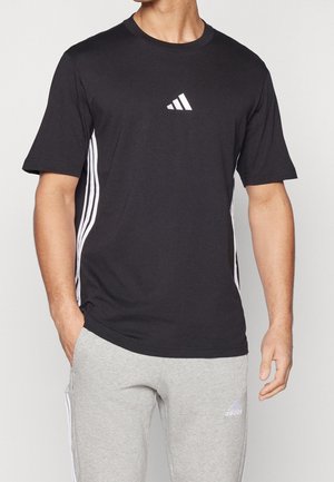 Mężczyzna noszący czarną koszulkę Adidas z białymi paskami po bokach oraz szare spodnie dresowe Adidas z logo na lewym udzie.
