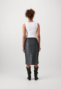 Osoba s kudrnatými vlasy má na sobě bílý top bez rukávů a černou koženou tužkovou sukni, stojí před jednobarevným světlým pozadím.