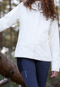 Veste imperméable blanche avec capuche, dotée de poches zippées et d'une texture lisse. Associée à un pantalon de plein air marine, mettant en avant un détail de logo.